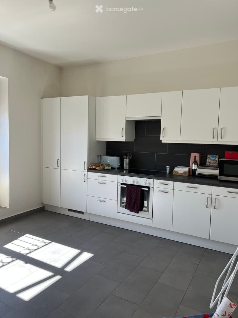 Modern kitchen with white cabinets, dark countertops, and stainless steel appliances including an oven, microwave, and toaster. A bottle and oven mitt are visible on the counter.