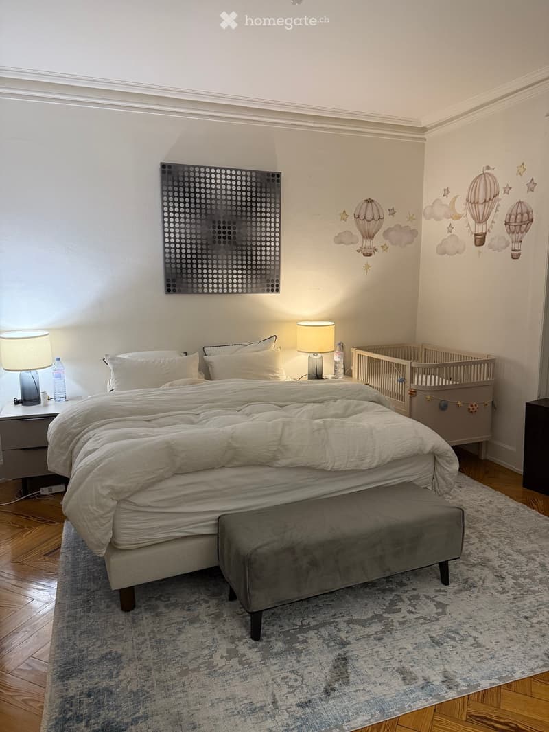 A well-lit bedroom with a large bed, nightstands, lamps, a grey ottoman, a crib, and decorative wall art featuring hot air balloons and stars.