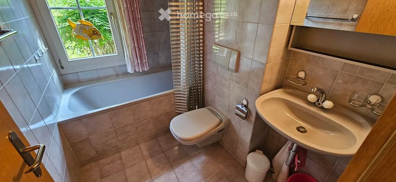 Bathroom with a bathtub, toilet, sink, mirror, and cabinet. The room features tiled walls and floor, a window with a curtain, and a small trash bin.
