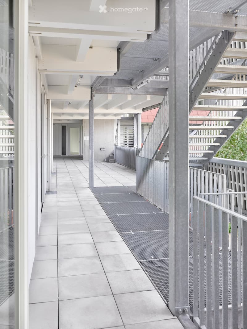 Modern, open-air corridor with concrete floors, metal railings, and stairs leading to an upper level. Features a glass wall on one side and a metal mesh walkway on the other.