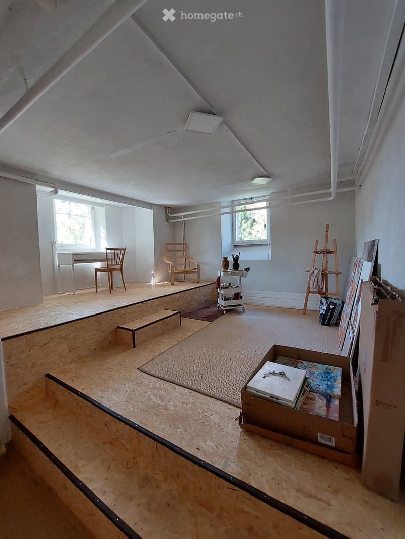 an empty room with wooden floor, white walls, two windows, two wooden chairs, a desk, some paintings and art supplies