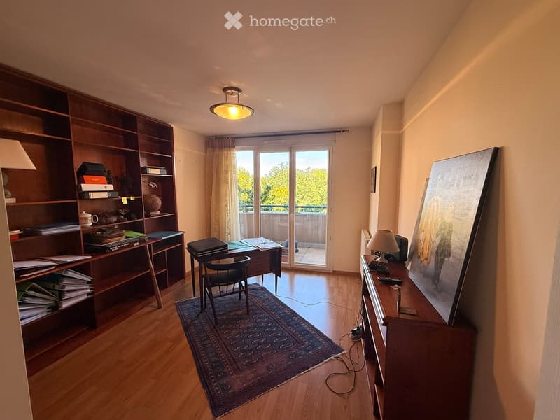 Spacious room with wooden flooring, large bookshelf, desk, chair, and artwork on the wall. Features a sliding glass door leading to a balcony.