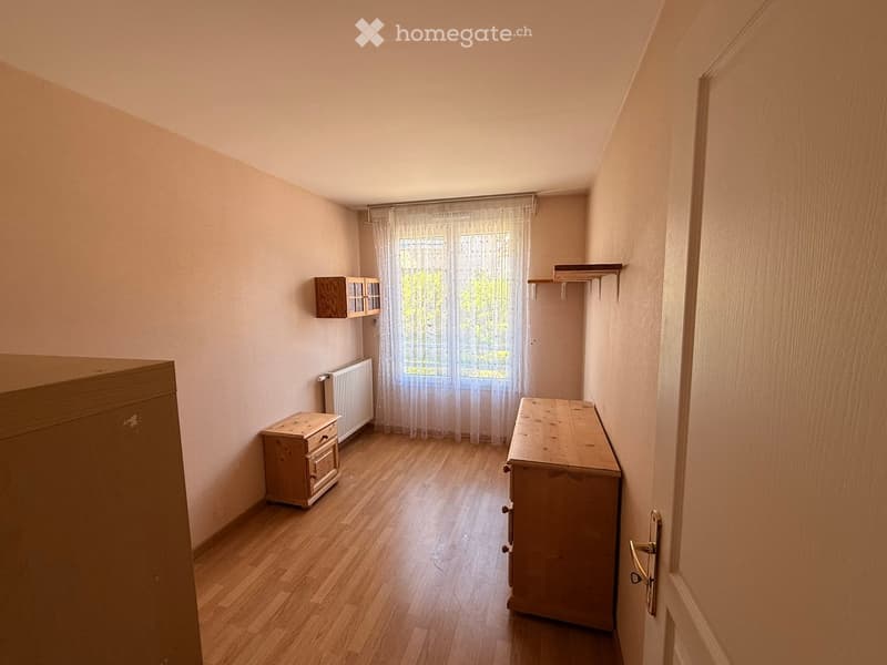 A small, unfurnished room with wooden flooring, a window with sheer curtains, a radiator, and a few wooden cabinets and shelves.