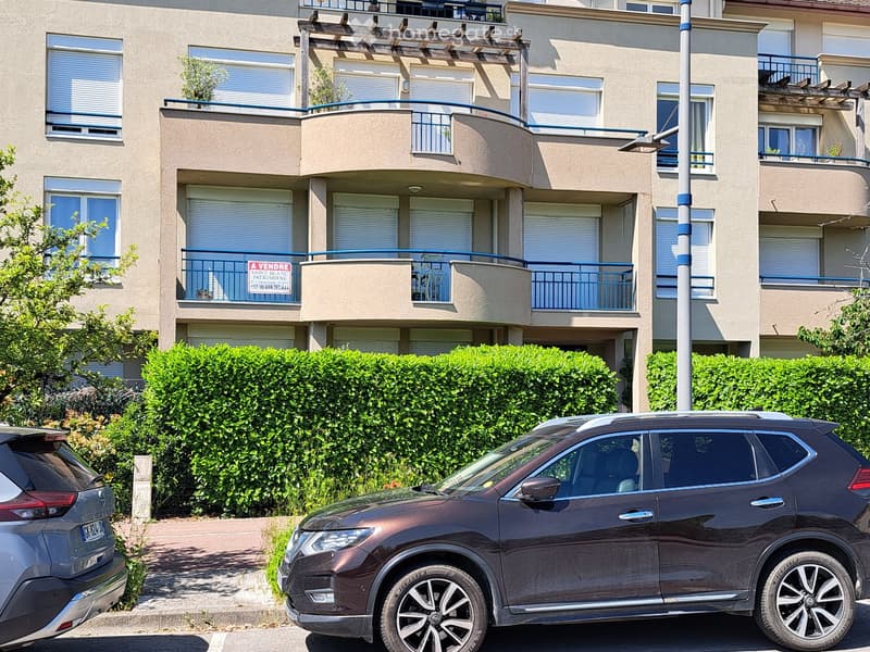A modern multi-story apartment building with balconies, large windows, and a sign indicating it is for sale. The building is surrounded by greenery and has parked cars in front.