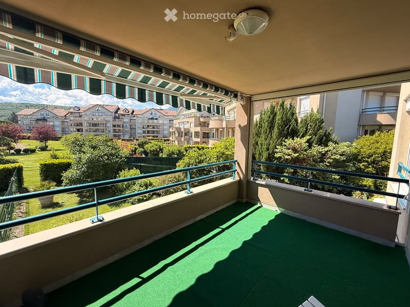 Balcony with green artificial turf flooring, metal railings, striped awning, and scenic view of residential area and greenery.