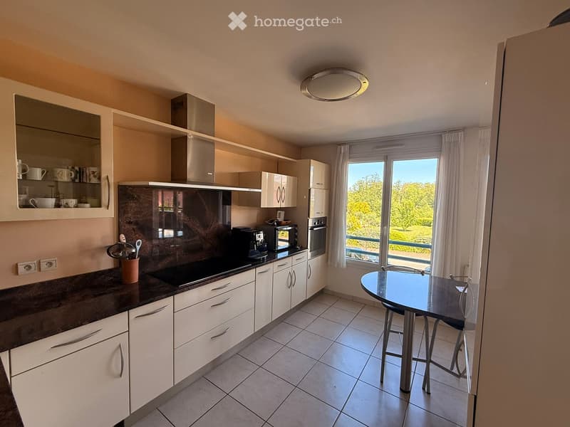 Modern kitchen with white cabinets, dark countertops, built-in oven, microwave, coffee maker, and a round dining table with stools. Large windows offer a scenic view of greenery.
