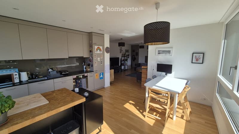 Modern kitchen with light-colored cabinets, stainless steel appliances, and a wooden island. A small dining area with a table and chairs is visible, along with a living area featuring a sofa and a TV.