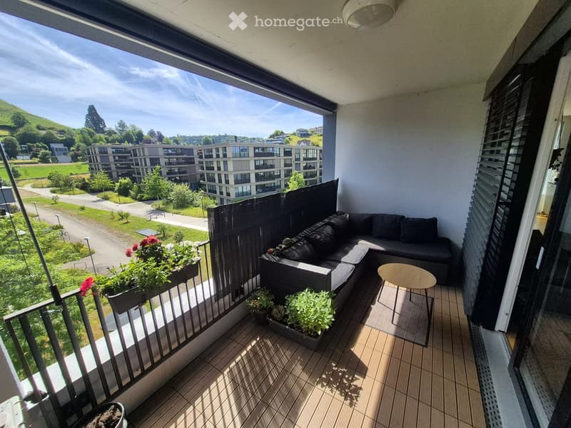 Balcony with a view of a green landscape and modern buildings, featuring a black sofa, a small round table, and several potted plants.