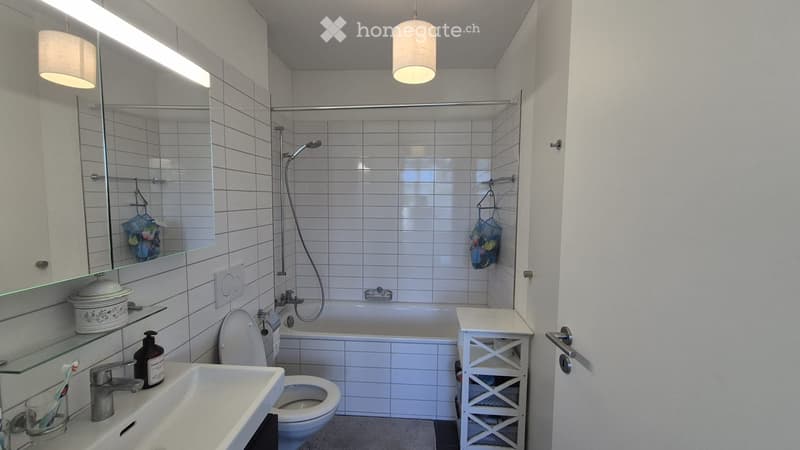 Modern bathroom with white tiles, a bathtub, toilet, sink, and a shower area. Includes a mirror, shelves, and a small storage unit.