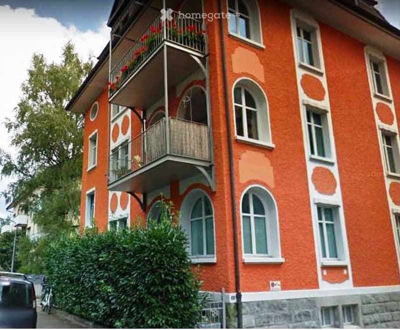 Apartment building with 3 stories, balcony on the first floor, brick and white exterior walls, large glass windows, small garden