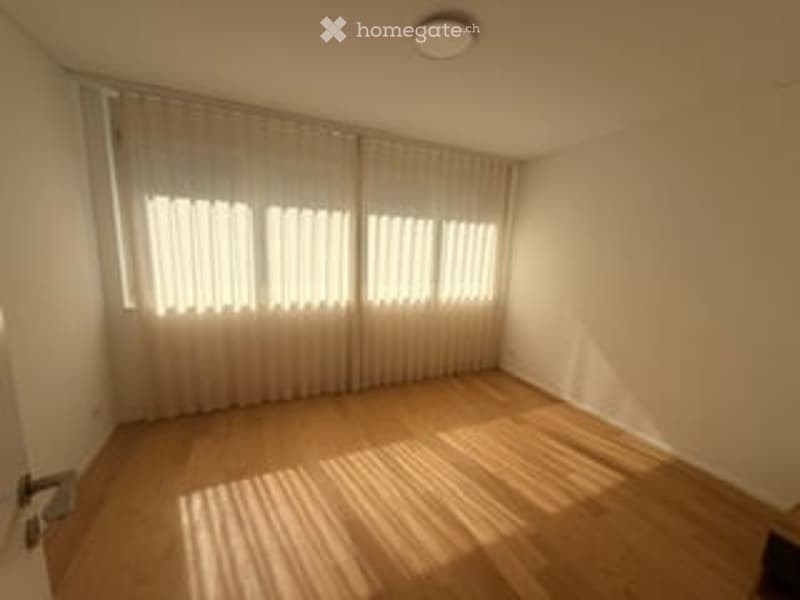 Empty room with wooden flooring, large windows with white curtains, and a ceiling light fixture.