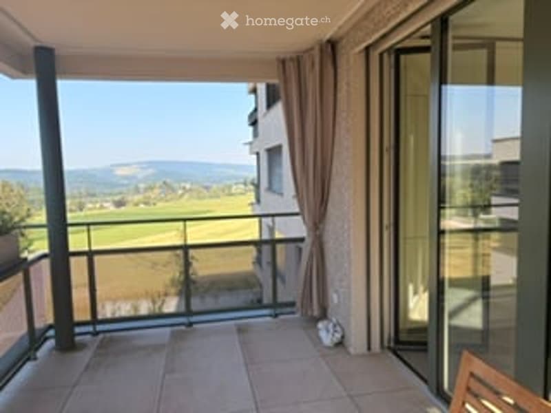 Balcony with a scenic view of a green field and distant mountains, equipped with a metal railing and large glass doors leading to the interior.