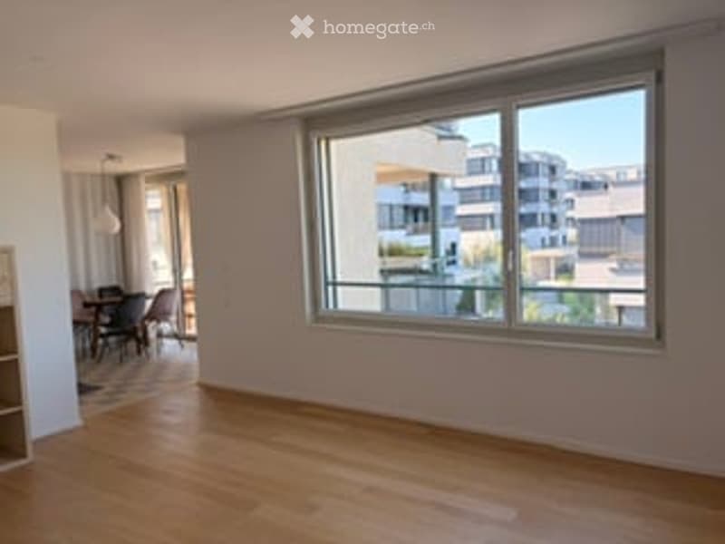 Empty room with wooden flooring, large window with a view of a cityscape, and a small dining area visible in the background.