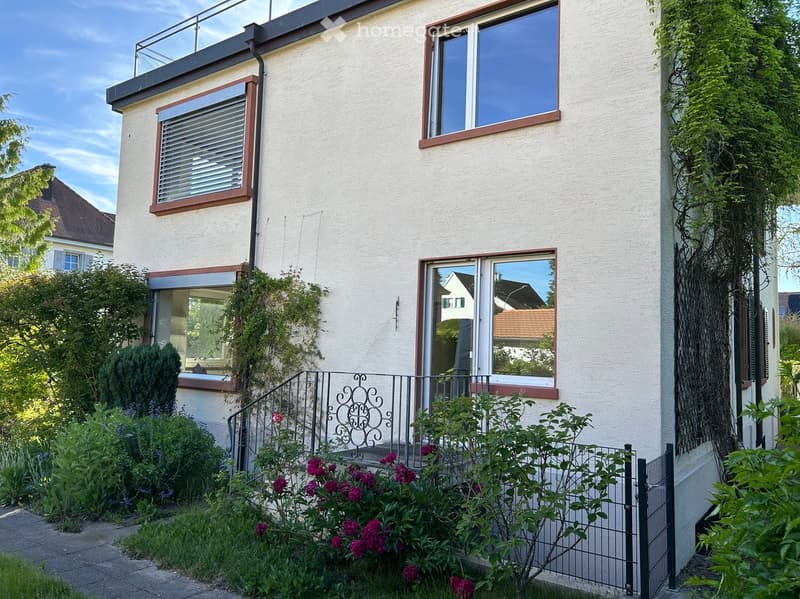 Two-story house with white exterior, multiple windows, and a small balcony on the roof. The front yard is landscaped with flowers, shrubs, and a decorative metal railing leading to the entrance.