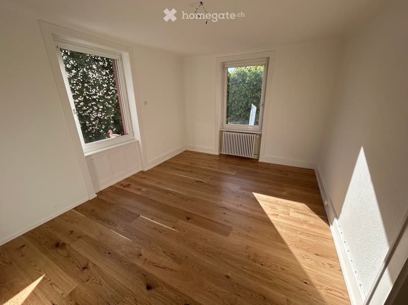 Empty room with light wooden flooring, white walls, and two windows showing greenery outside. No furniture or equipment present.