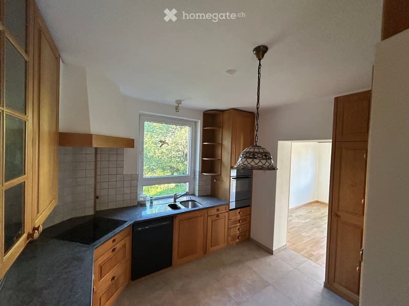 Bright kitchen with wooden cabinets, a sink, oven, dishwasher, and pendant light. A window offers a view of greenery outside.