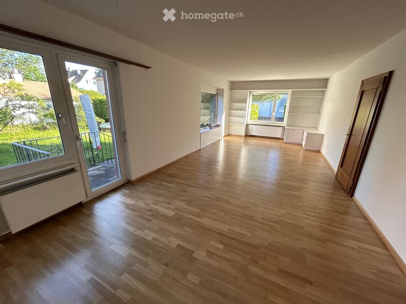 Empty room with wooden flooring, white walls, built-in shelving, and large windows with a view of greenery outside.