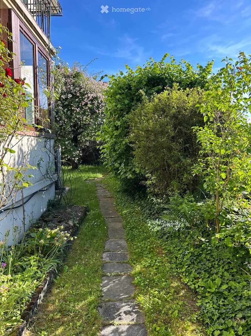 A narrow garden pathway lined with lush greenery and flowering bushes leads to a house with a balcony and large windows.