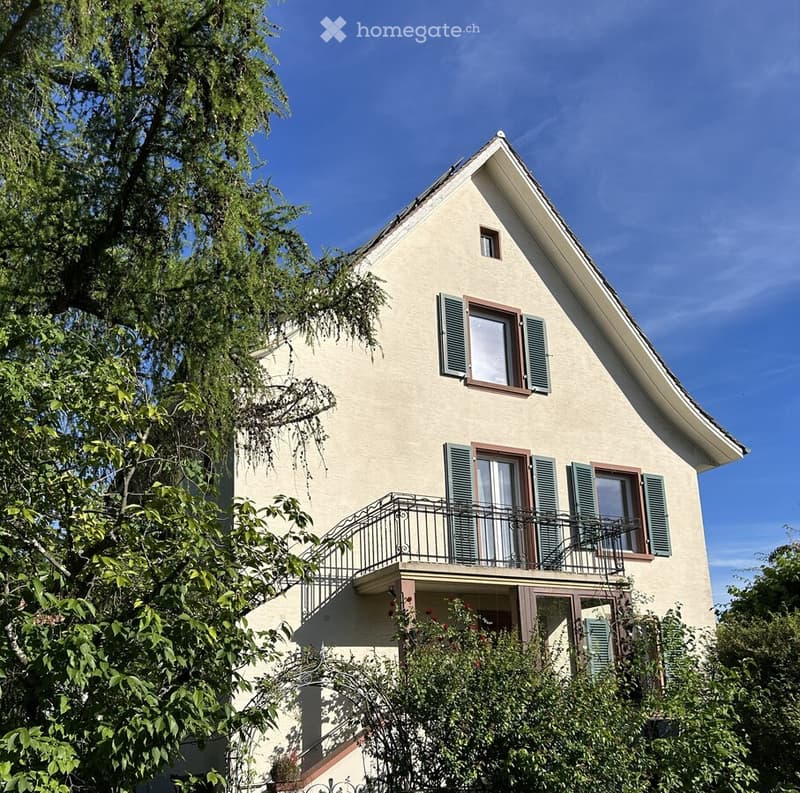 Two-story house with beige walls, green shutters, and a balcony on the first floor.