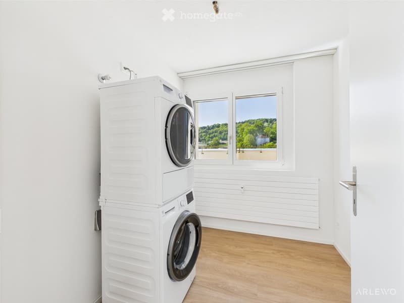 Bright, modern laundry room with white walls, wooden flooring, and a stacked washer and dryer unit. Large windows offer a view of greenery outside.