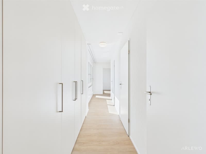 A clean, modern hallway with white walls, light wooden flooring, and recessed ceiling lighting. The hallway features several closed doors with sleek metallic handles, leading to other rooms.
