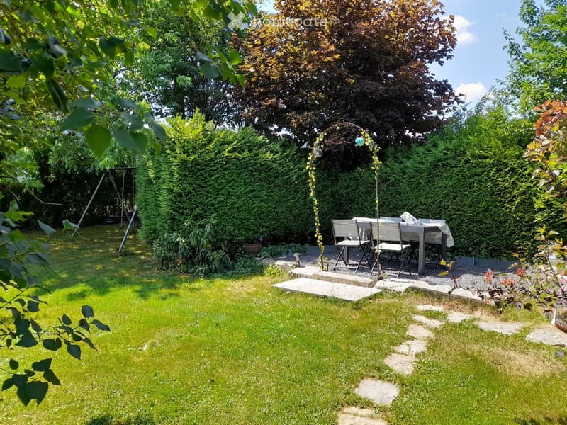 Well-maintained garden with a lawn, a small seating area with a table and chairs, a swing, and a decorative archway. The area is surrounded by lush greenery and trees.