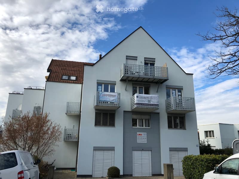 Modern two-story building with white exterior, multiple balconies, and a tiled roof. Features include large windows, a garage entrance, and signage for "Shkodra Bau AG" and "LMS Immobilien-Service".