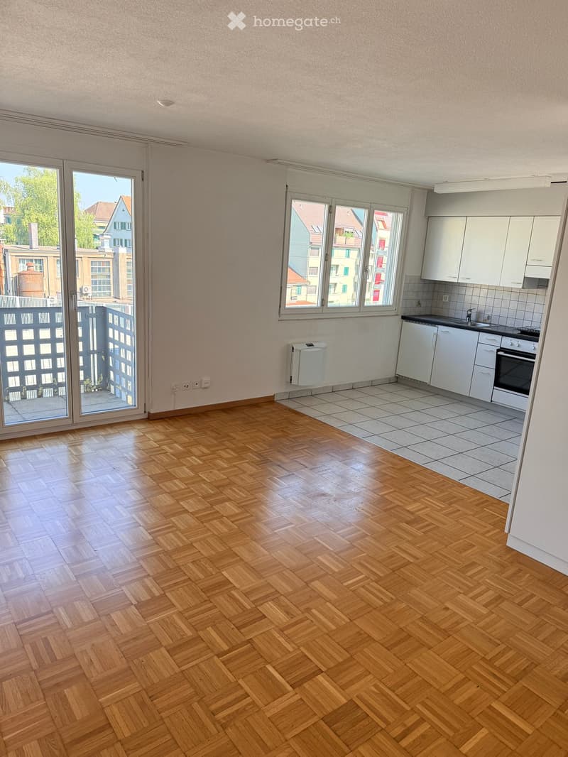 Open-plan living and kitchen area with wooden herringbone flooring, white kitchen cabinets, sink, oven, and radiator.