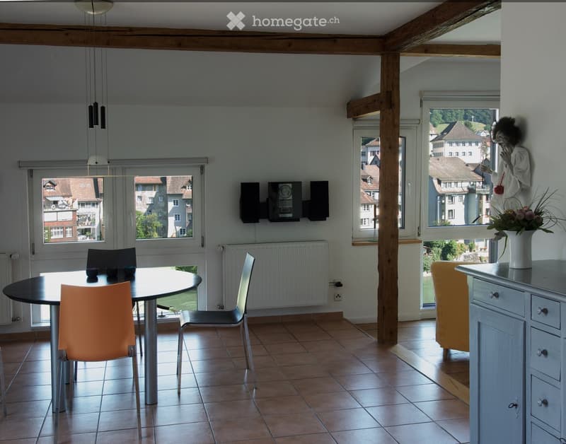 Bright dining area with a round black table, four chairs, a vase with flowers, and a radiator. Large windows offer a view of the town.
