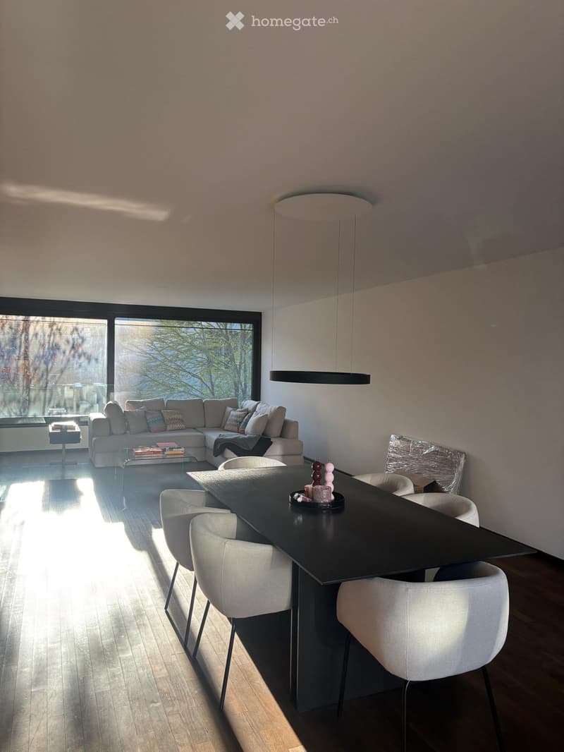 Modern living and dining area with a sectional sofa, coffee table, dining table, and chairs. Large windows allow natural light to fill the space.