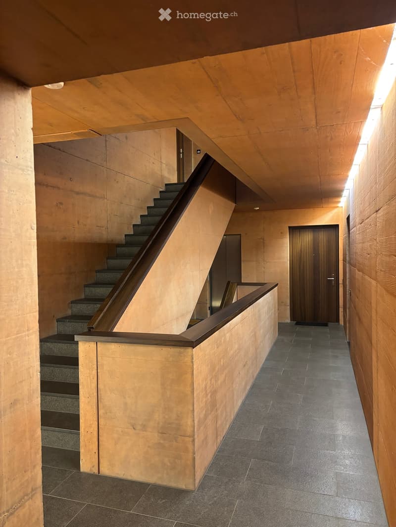 Modern hallway with a staircase, wooden railing, and a door at the end. The walls are light brown, and the floor is tiled in dark gray.
