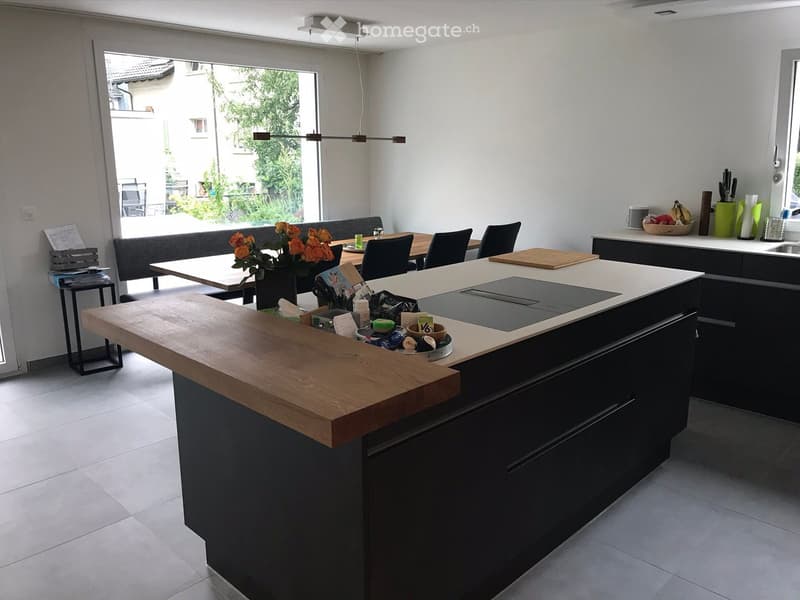 Modern kitchen with island, stove, sink, and seating area. Features dark cabinets, light countertops, and a dining table with chairs.