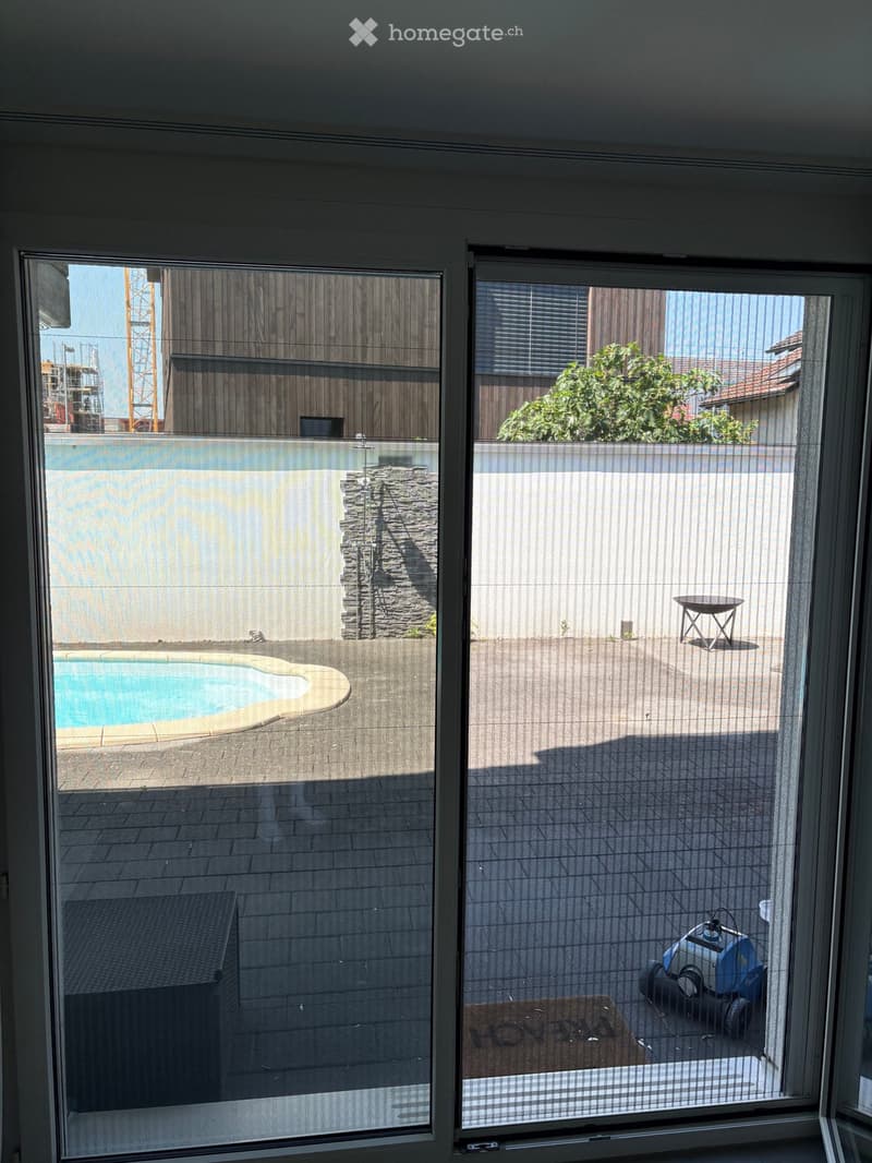 View from inside through large glass doors showing a swimming pool, patio area, and outdoor furniture.