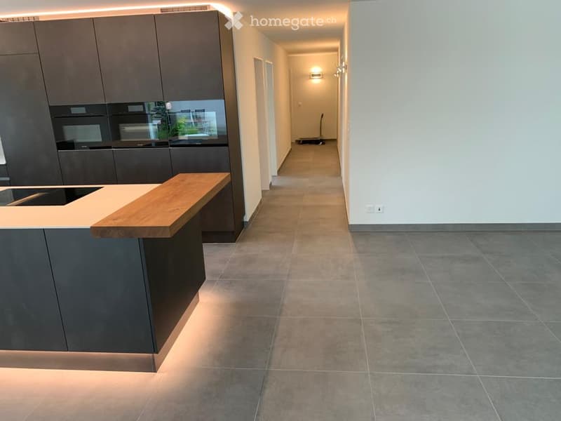 Modern hallway with tiled floor, grey walls, and a kitchen area visible on the left with dark cabinets and a wooden countertop.