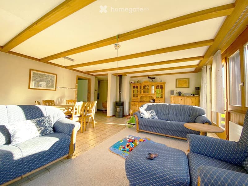 Bright living room with exposed wooden beams, blue patterned sofa, ottoman, armchair, coffee table, dining area with chairs, and a cabinet with glass doors.