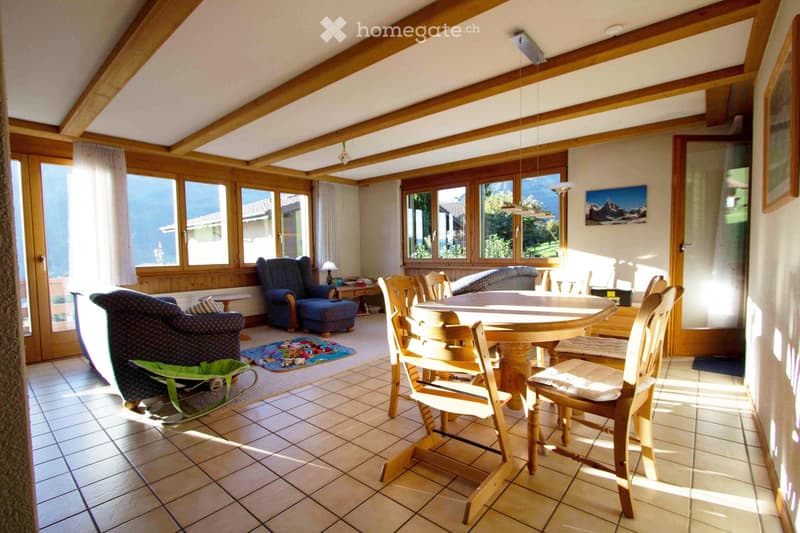 Bright living room with large windows, wooden beams, tiled floor, sofa, chairs, dining table, and artwork.
