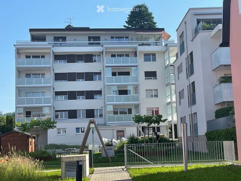 Modern multi-story residential building with balconies, large windows, and a landscaped garden area with a playground and seating.