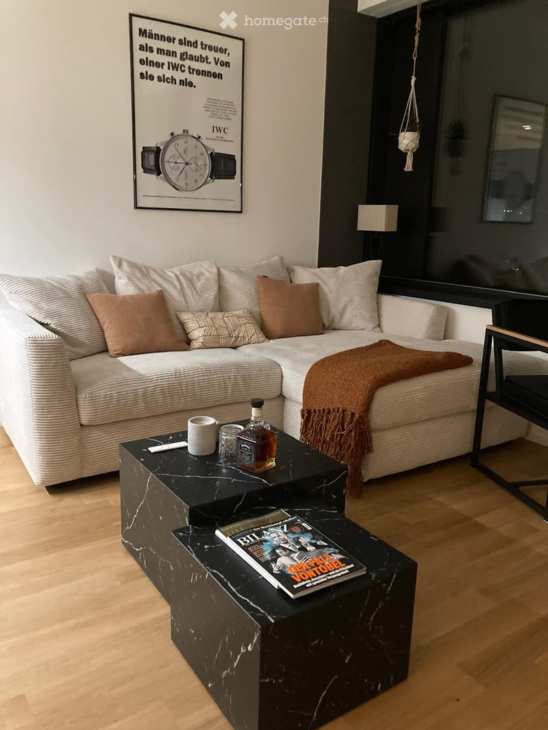 Modern living room with a white sofa, black marble coffee table, throw blanket, and a magazine on the table. The room features wooden flooring and a large window with a view.