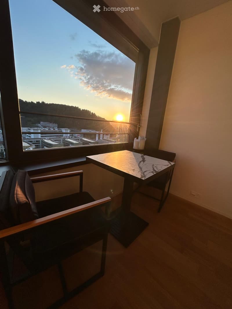 A small, modern room with a large window offering a view of the sunset, featuring a wooden chair, a marble-top table, and a pair of candles on a shelf.