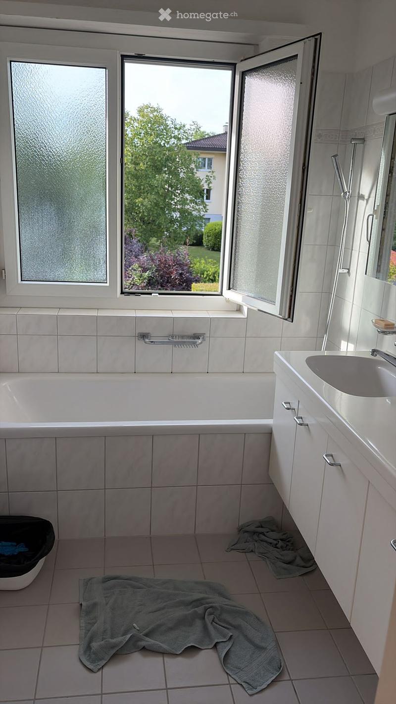 Bathroom with a bathtub, sink, and mirror. The room has tiled walls and floor, with a window offering a view of the garden. Towels are placed on the floor, and a trash bin is visible.