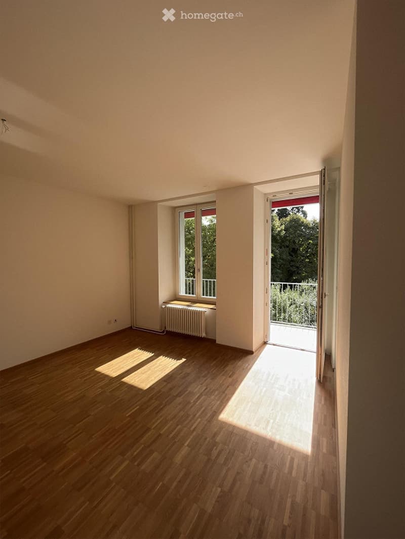 Empty room with wooden flooring, white walls, a window with a radiator, and an open sliding door leading to a balcony.