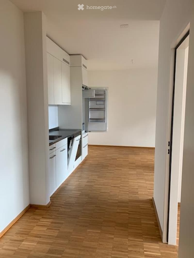 Empty kitchen with white cabinets, wooden flooring, and an open refrigerator. No furniture or appliances are present.