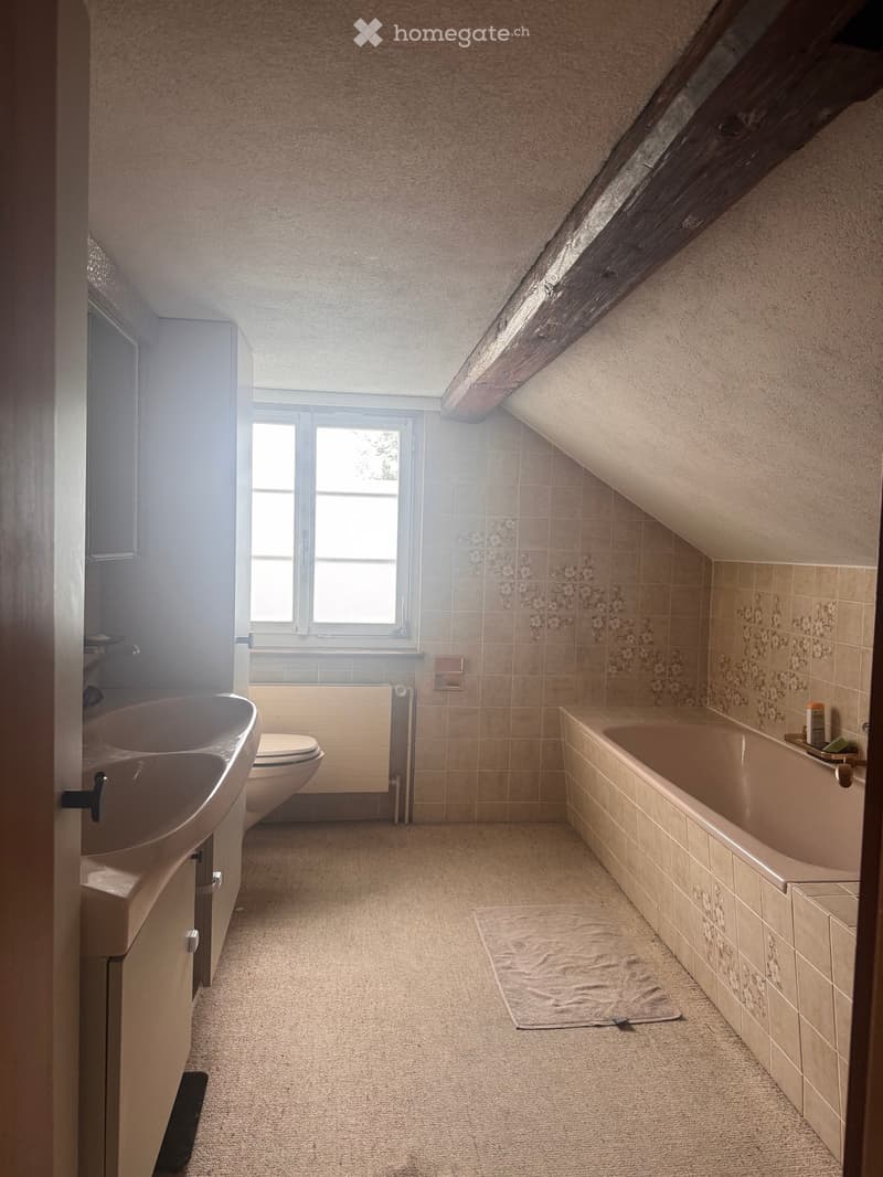 Bathroom with a sink, toilet, bathtub, and a towel on the floor. The room has beige tiles and a slanted wooden ceiling.