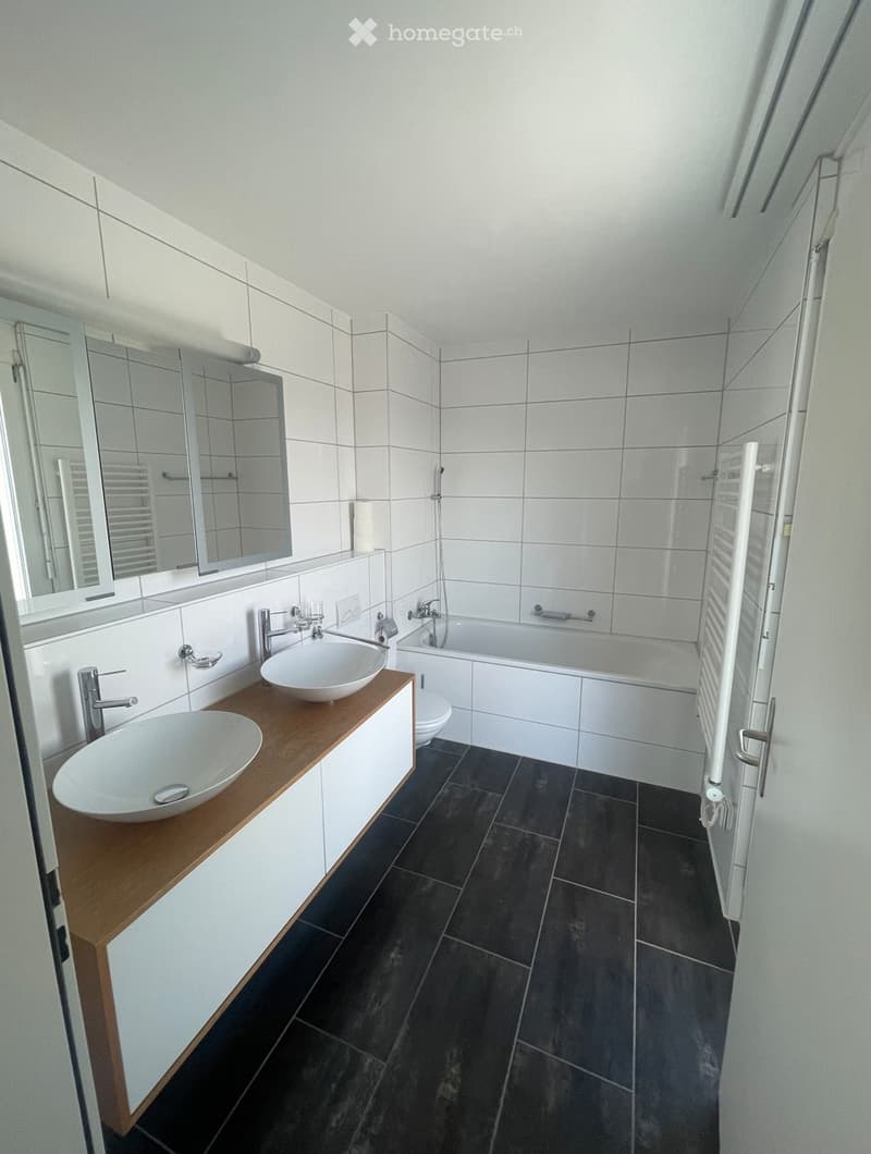 Modern bathroom with white tiles, dark floor, double sink vanity, bathtub, and large mirror.