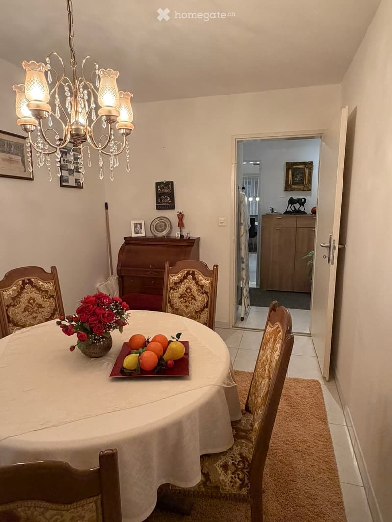 Elegant dining room with a round table covered in a white tablecloth, surrounded by four ornate wooden chairs. A chandelier hangs from the ceiling, and a small cabinet with decorative items is visible in the background. A doorway leads to another room wit