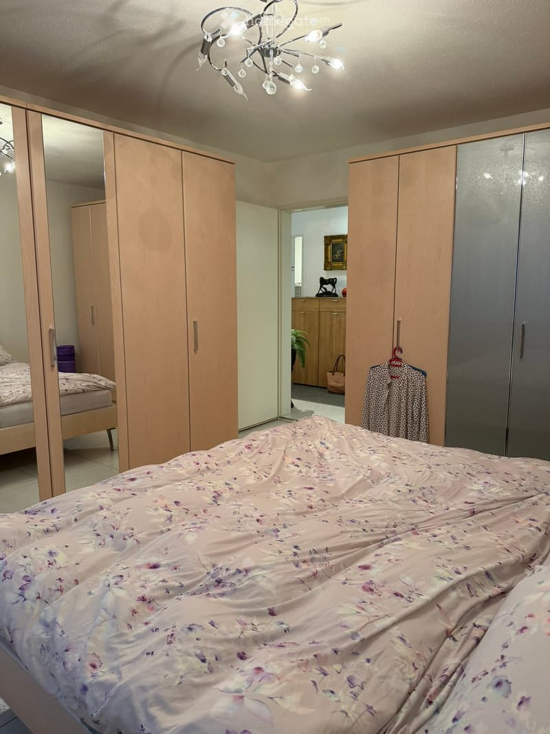 Well-lit bedroom with a large bed covered in a floral duvet, a wooden wardrobe, a mirrored closet, and a decorative chandelier.