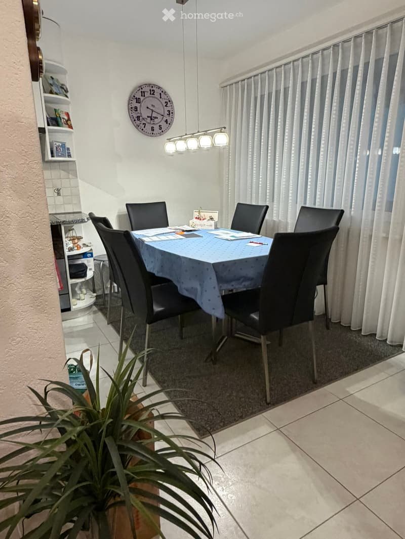 Dining area with a round table covered in a blue tablecloth, surrounded by six black chairs. A wall-mounted clock is above the table, and a shelf with decorative items is visible to the left. A large window with white curtains allows natural light to fill