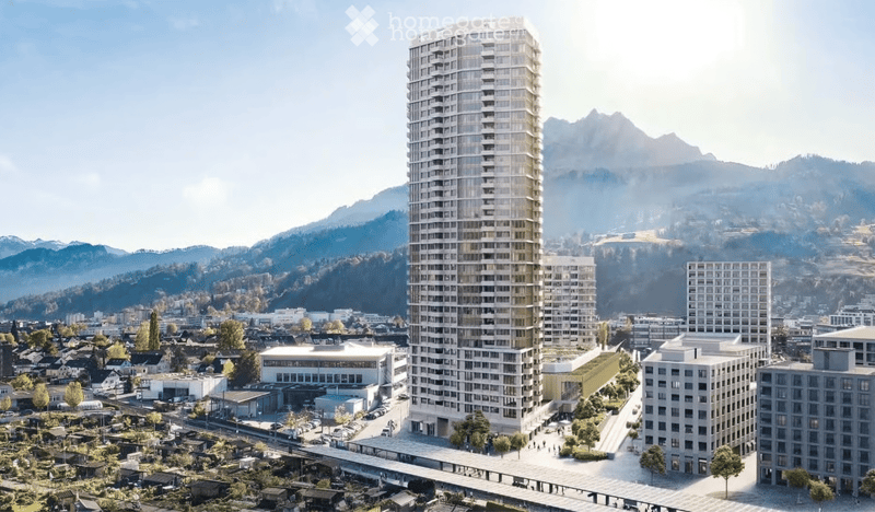 A modern high-rise building stands prominently in the foreground, surrounded by smaller structures and a scenic mountain backdrop.