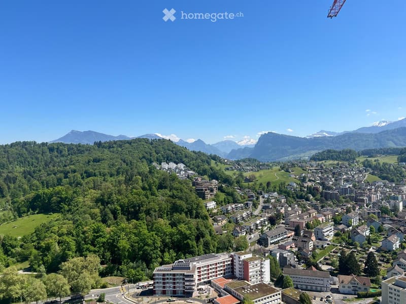 Aerial view of a town nestled in a valley surrounded by lush green forests and mountains, with a mix of residential and commercial buildings, roads, and a clear blue sky.