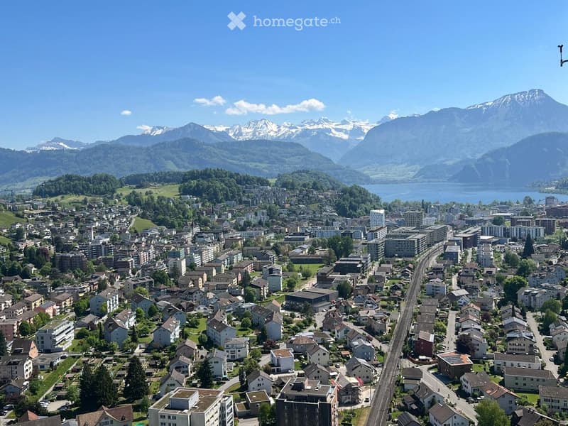 Aerial view of a town nestled in a valley with surrounding mountains and a lake in the background. The town features a mix of residential and commercial buildings, roads, and green spaces.